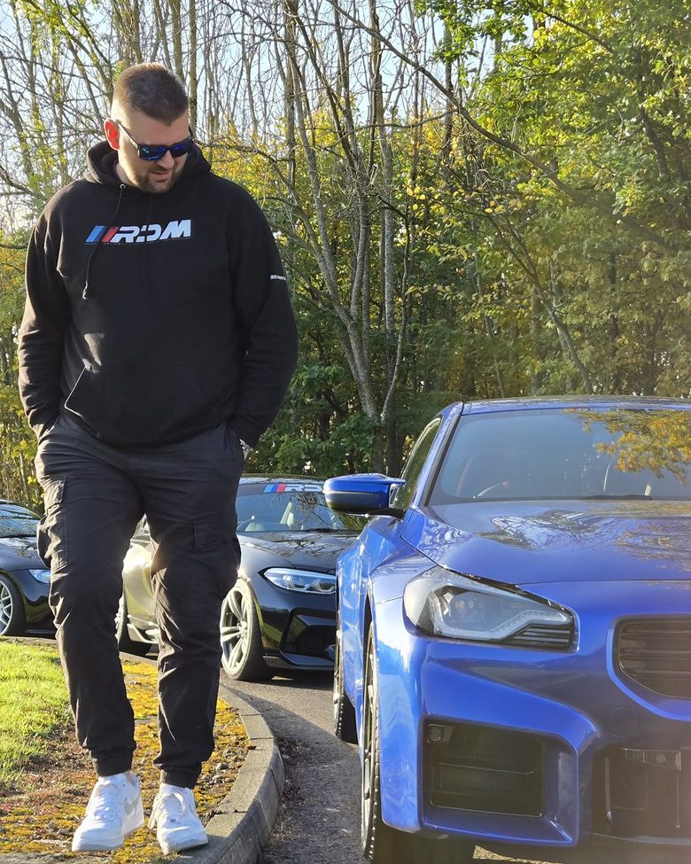 Robbie in an RDM hoodie, walking past a blue G87 M2